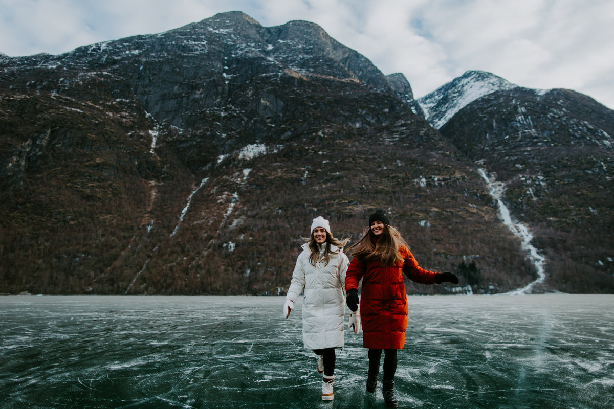 To kvinner i vinterjakker på frossen fjord med snødekte fjell i bakgrunnen